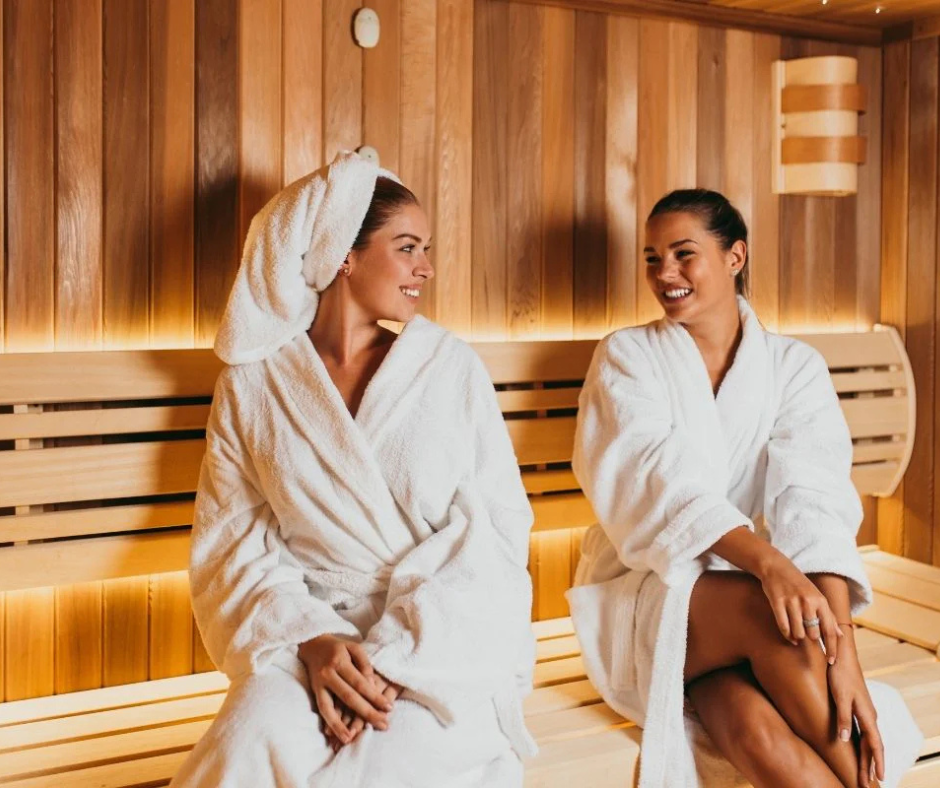 two ladies relaxing in a sauna, wearing white robes and smiling at one another.