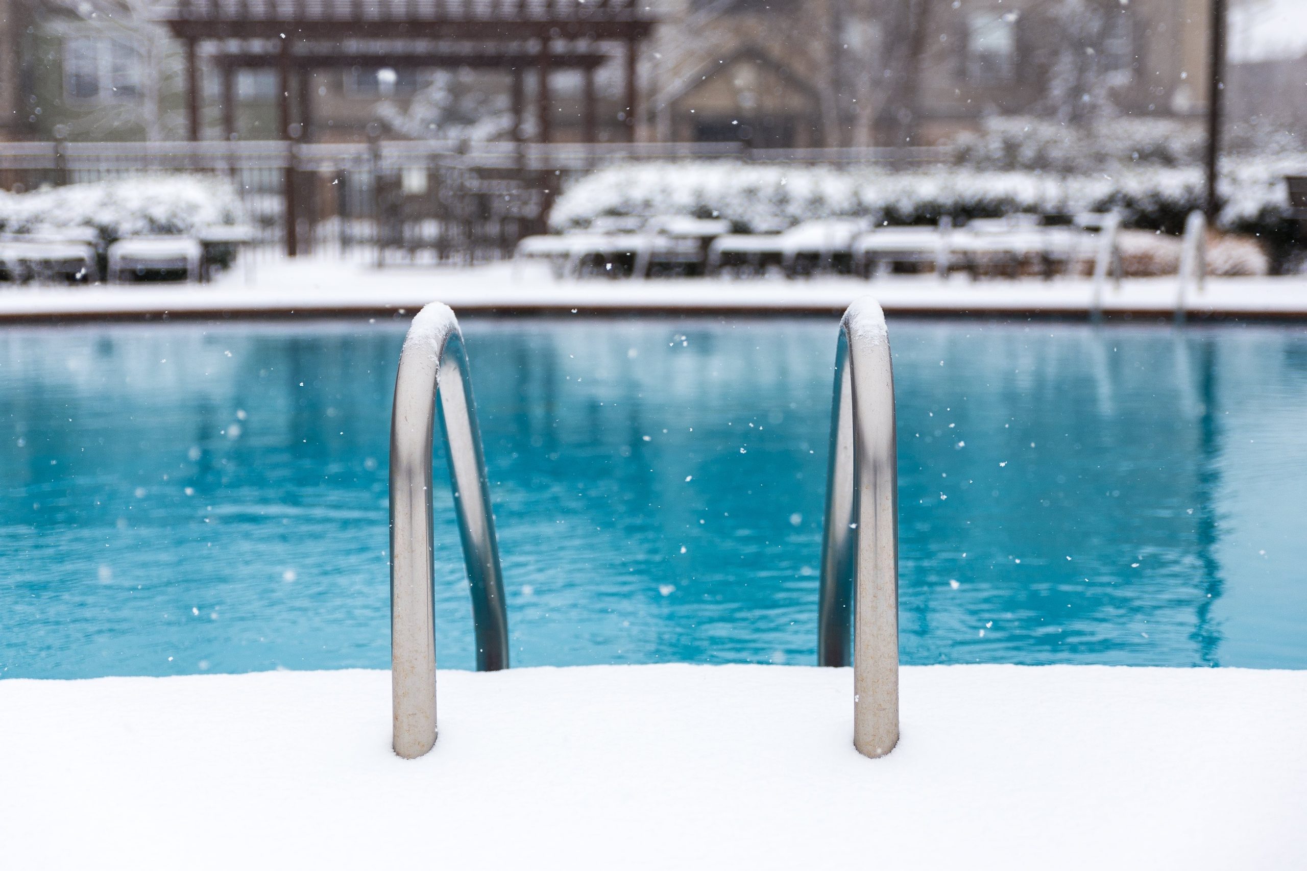 a close up photo from the point of view of someone standing at an inground pool ladder, facing out towards the water. there is snow falling as well as on the ground in the background. the ladder is in focus, while the pool and the background are a little blurry.