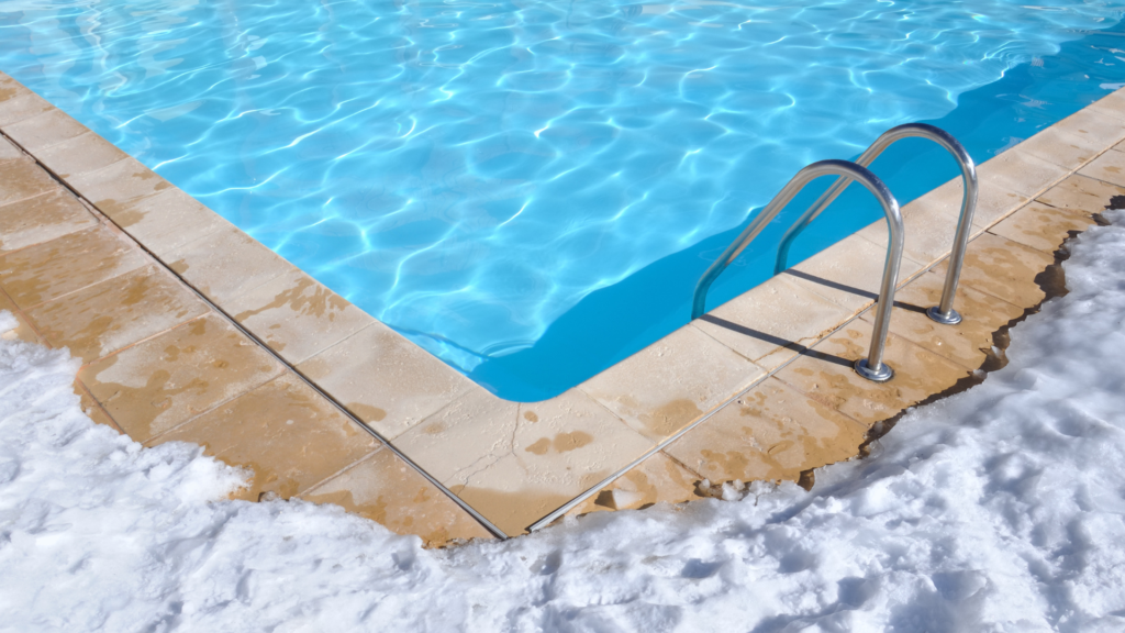 a close of photo of the corner of an inground pool. there is snow on the concrete surrounding it, and you can see the silver handrails of a ladder going into the water