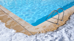 A close of photo of the corner of an inground pool. There is snow on the concrete surrounding it, and you can see the silver handrails of a ladder going into the water