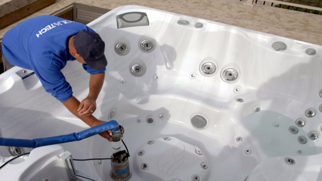 an overhead shot of an aqua-tech service technician using a submersible pump to vacuum water out of the footwell of a sundance spa. her is wearing a royal blue aqua-tech branded crew neck sweater, and a navy blue baseball cap. there is very little water left inside the tub, only in one of the seats on the right hand side of the photo. the interior acrylic of the tub is white. the exterior is not visible in the photo.