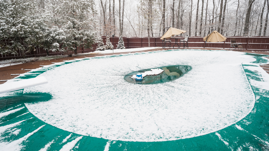 a photo of an inground pool covered by a turquoise pool cover, with snow on top. there is a reddish brown fence in the background with a pew patio tables with umbrellas. there are snowy pine trees on the left side, outside of the fenced in yard.