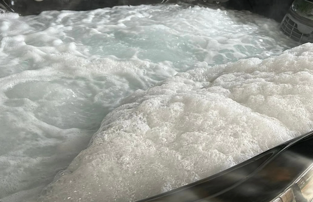 a close up photo of very foamy hot tub water. there are thick white bubbles on the surface of the water, and in the bottom right corner of the image the dark marbled acrylic of the tub is visible.