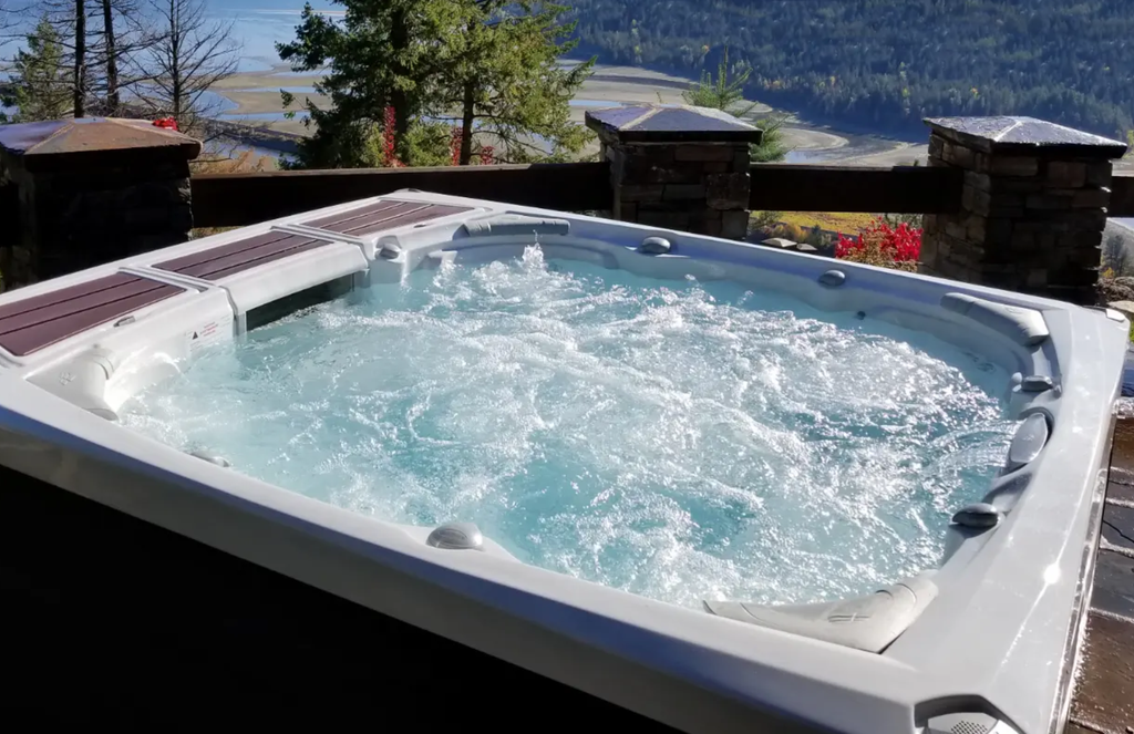 a photo of a hot tub with the cover off. th jets are on and the water is rippling and bubbling. there is a fence in the background with brick/stone pillars and wooden planks. there are trees and a landscape in the far background.