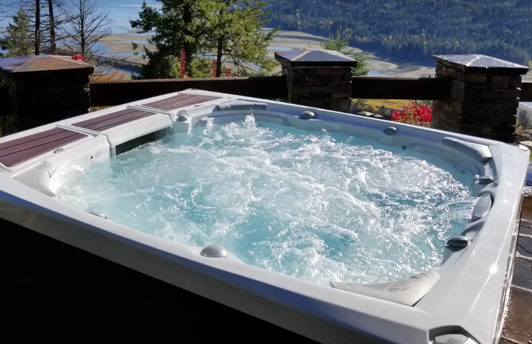 A photo of a hot tub with the cover off. Th jets are on and the water is rippling and bubbling. There is a fence in the background with brick/stone pillars and wooden planks. There are trees and a landscape in the far background.