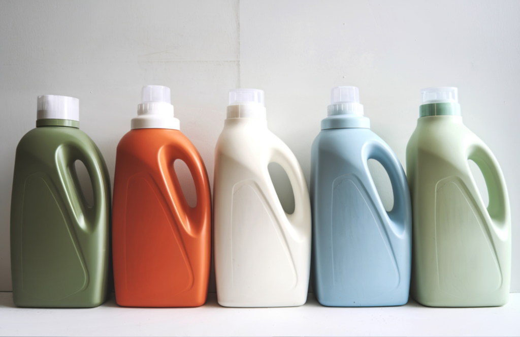 a photo with a light gray background and a row of various coloured detergent bottles. from left to right; dark green, orange, white, pale blue, and sage green.