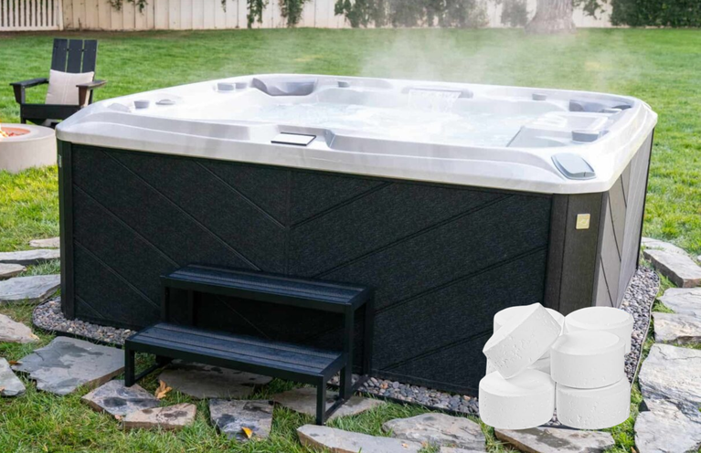 A photo of a hot tub in a yard with green grass. There are grey and brown patio stones beneath the spa and surrounding it, and a set of dark steps leading up to the hot tub. There is a pile of sanitizer tablets in the right side of the picture.
